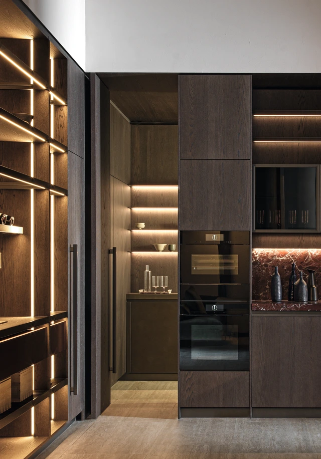 Stacked built-in ovens in tall dark wood kitchen cabinetry beside illuminated shelves and a burgundy veined splashback.