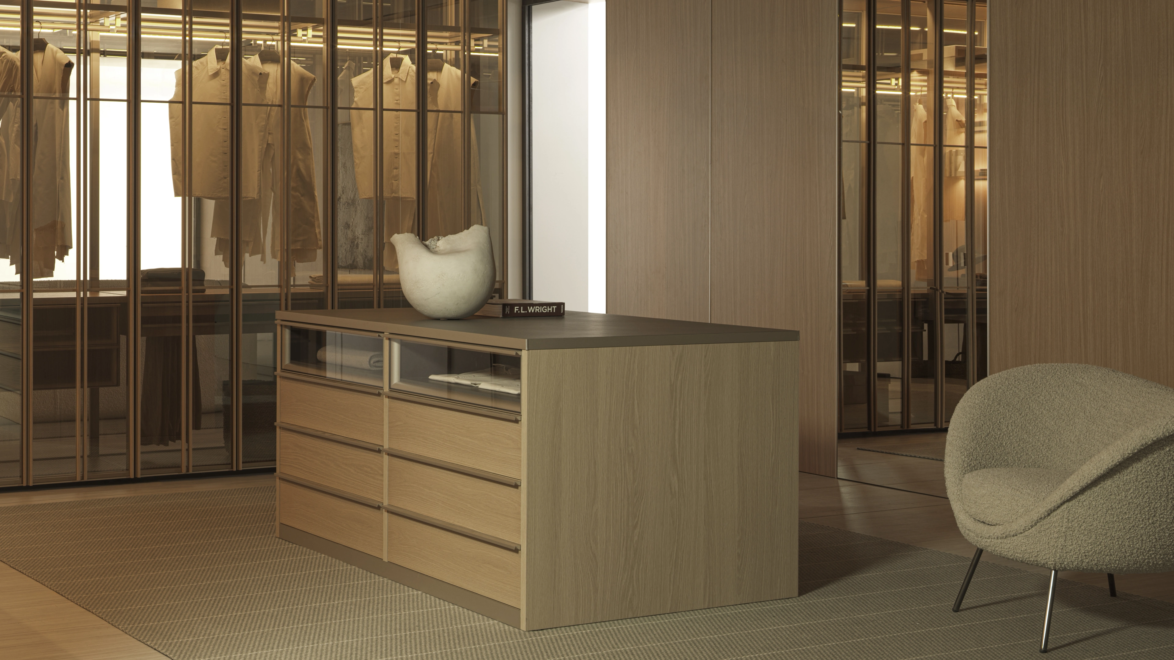 Walk-in closet with illuminated glass wardrobes, a light wood drawer island and a cream textured accent chair.