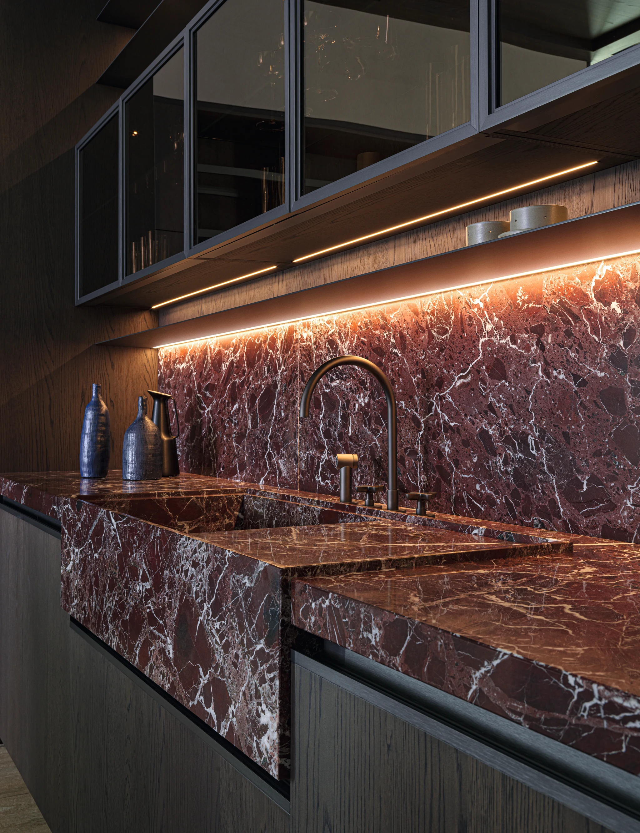 Burgundy veined sink and matching splashback beneath dark glass-front kitchen cabinets with warm strip lighting.