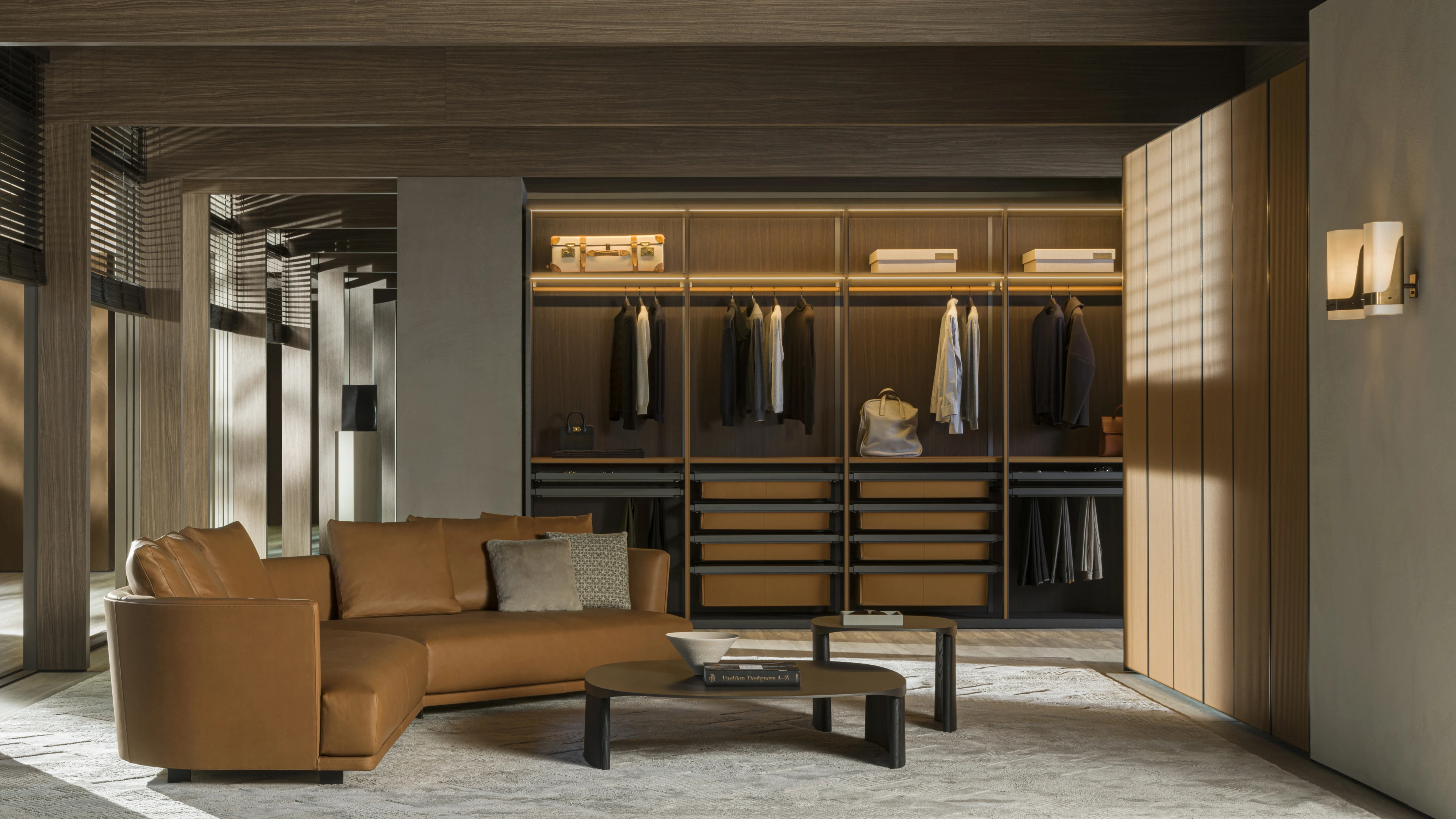 Open wood closet with illuminated hanging rails and tan drawers behind a curved tan sofa and low coffee tables.